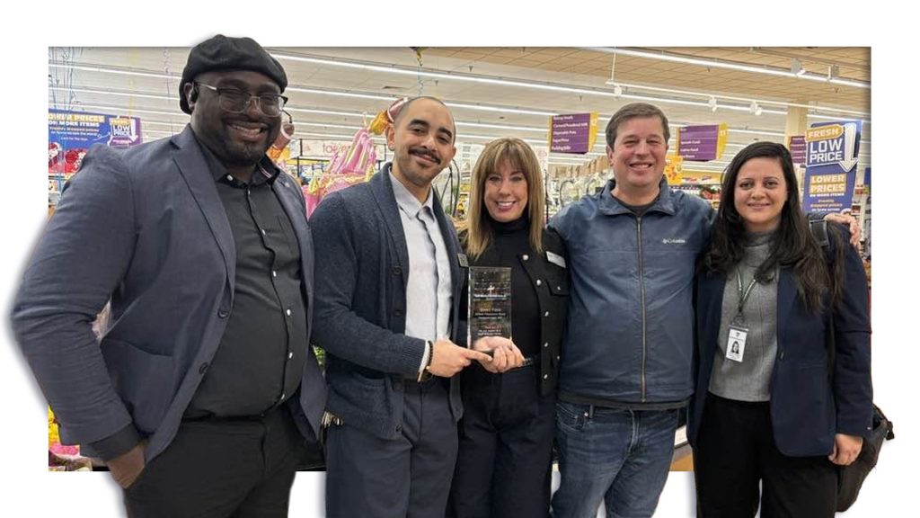 Five people smile together in a grocery store while presenting a community partnership award, with one person holding a recognition plaque.