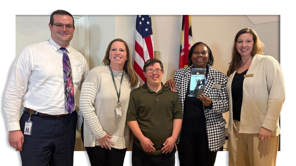 Five people pose together at an award presentation, including a participant standing with staff and a presenter holding a recognition plaque in front of the American and Maryland flags.