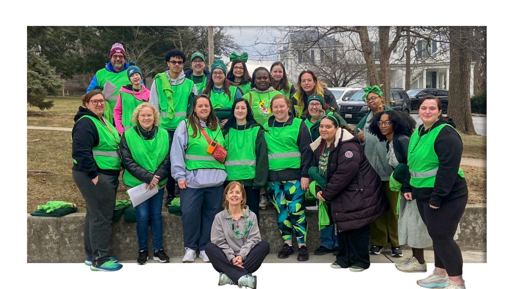A group of participants and staff wearing bright green safety vests pose together outdoors during a community volunteer event, smiling for a group photo.